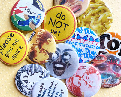 I frequently make new button designs! Here, we have several designs. Two yellow buttons, each saying "please give me space" or "do NOT pet", dum-dum wrappers, top ramen wrappers, collaged faces, and lesbian-themed buttons saying "studtastic" and "girlkisse