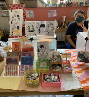 An array of products displayed on a location-provided table, about 6 ft. in length. This shot focuses on the viewer's left side of the table. It features my home-printed zines, linocut cards, and buttons. My fiance is seated in the back with a mask and pea